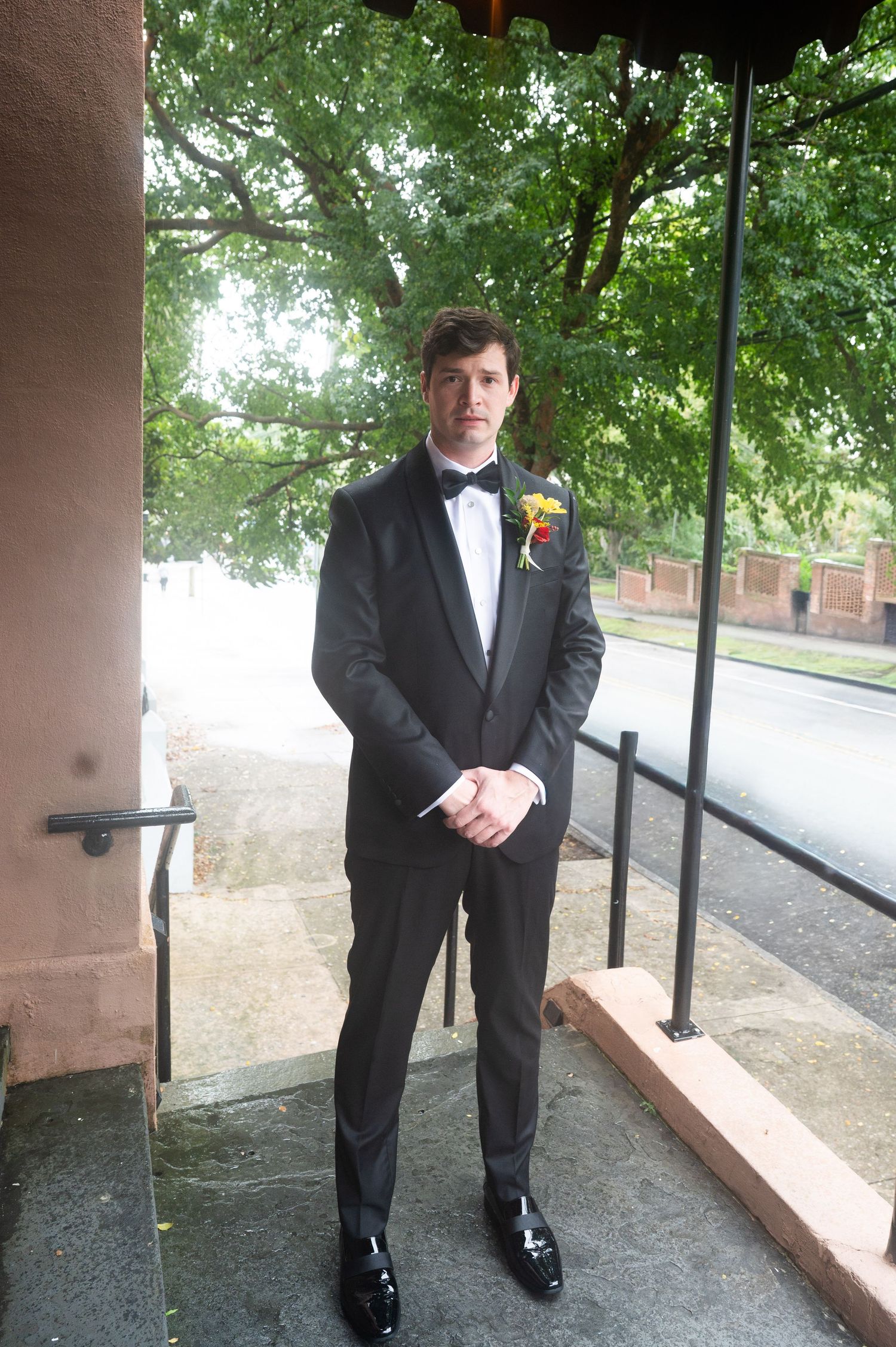 A person in a formal black tuxedo and bow tie stands on a covered outdoor porch area.