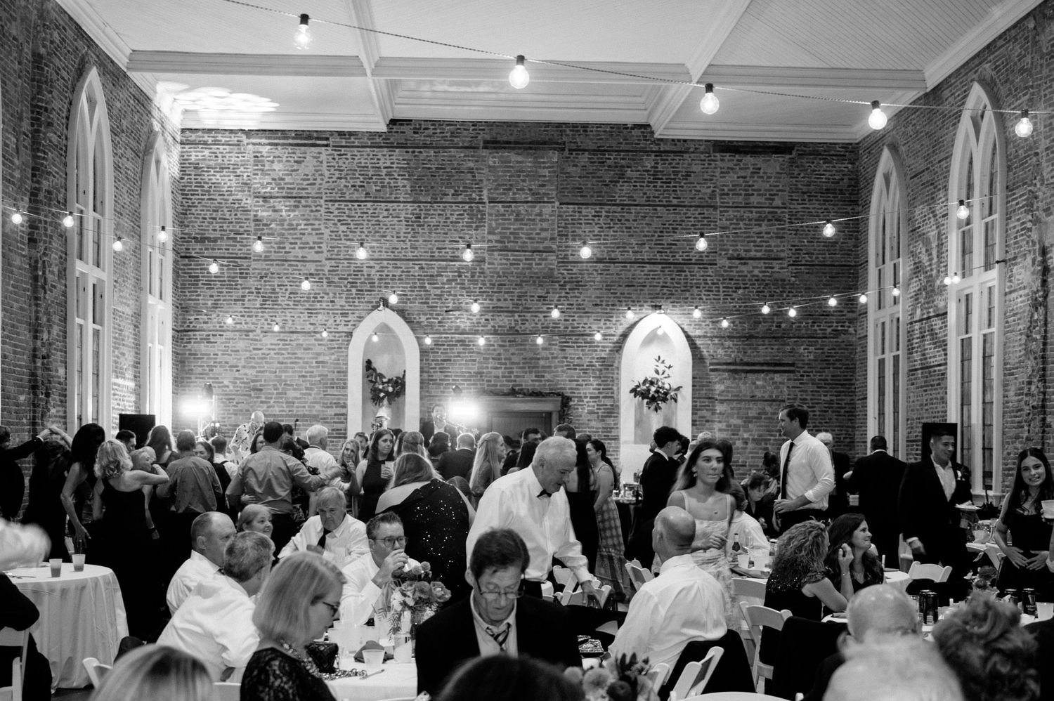 Wedding guests mingle in an industrial-style reception venue with exposed brick walls and string lights overhead.