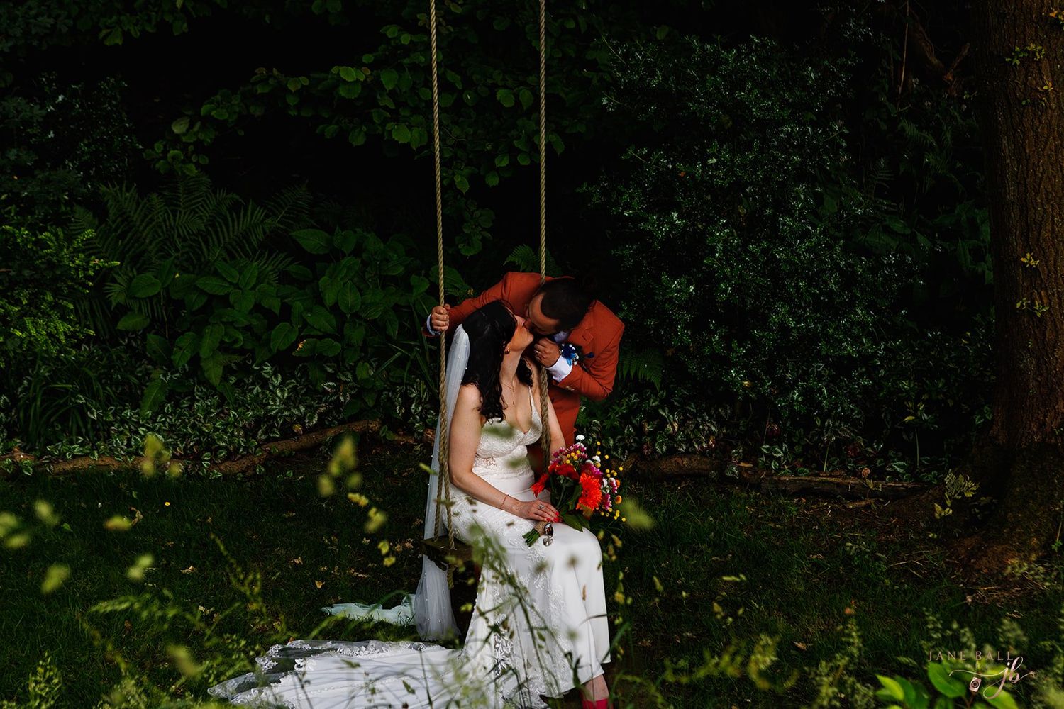 Bride and groom sharing a romantic kiss on a wooden swing surrounded by lush green forest trees. Hilltop Country House