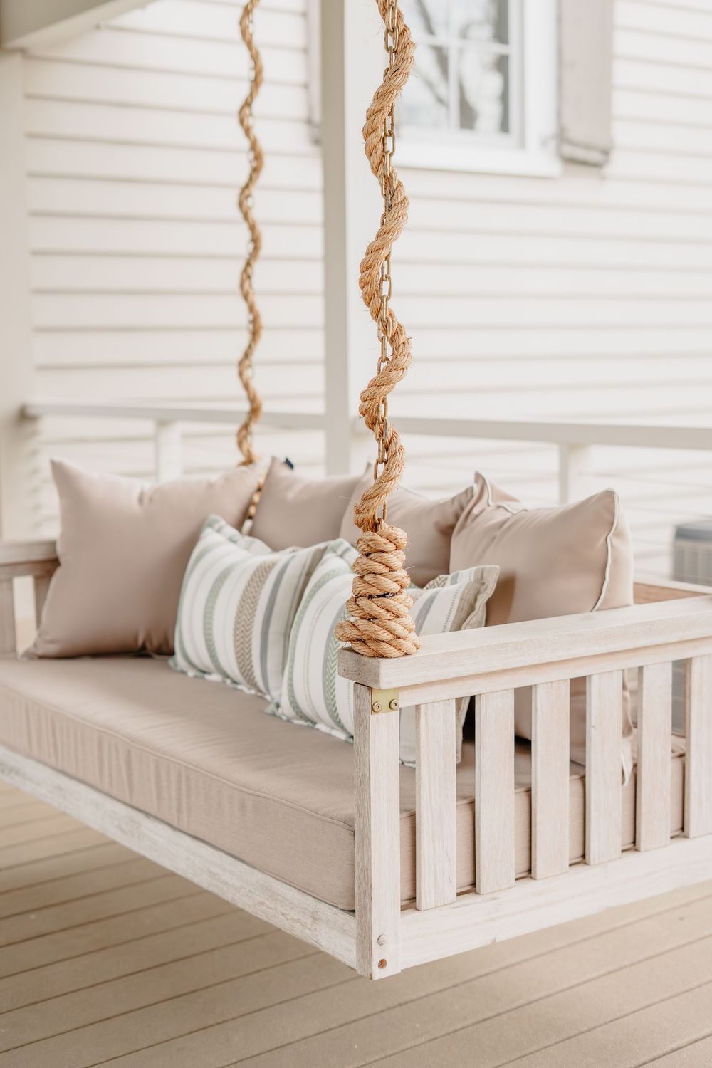 White wooden porch swing with neutral beige cushions and striped pillow, suspended by twisted rope on a bright veranda.