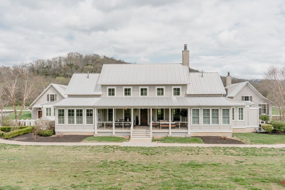 Large white modern farmhouse with metal roof, wraparound porch, and scenic mountain backdrop.