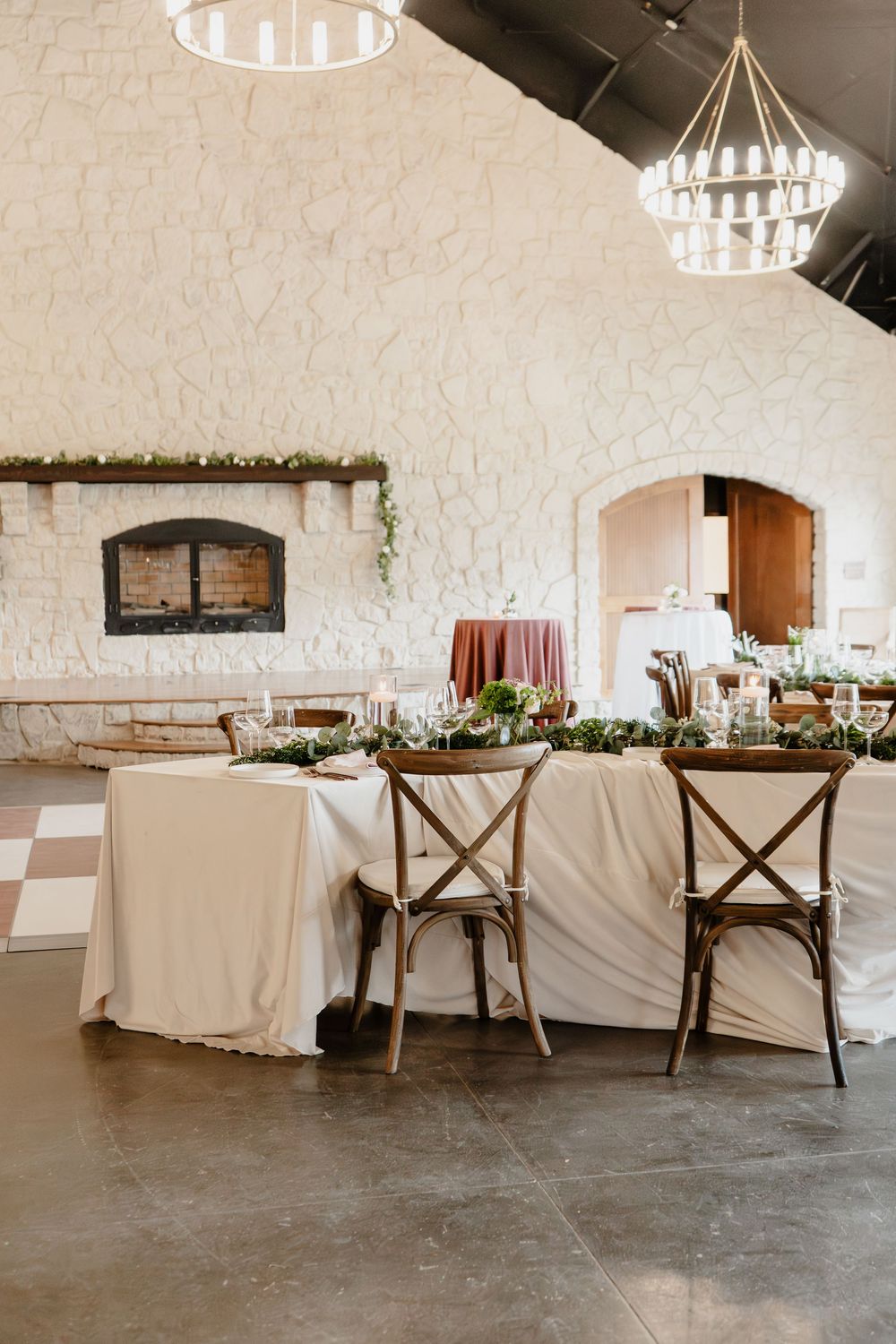 Elegant wedding reception table with white linen, greenery runner, candles, and cross-back chairs in a stone venue.
