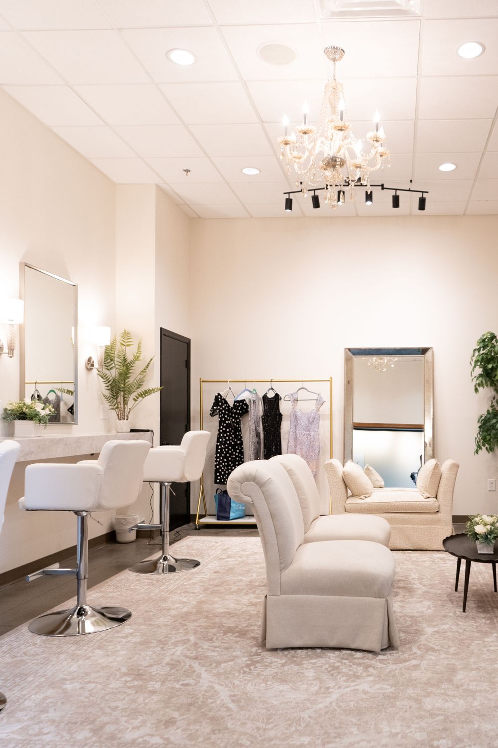 Elegant bridal suite with white tufted sofa, styling chairs, large mirror, chandelier, and clothing rack with dresses.