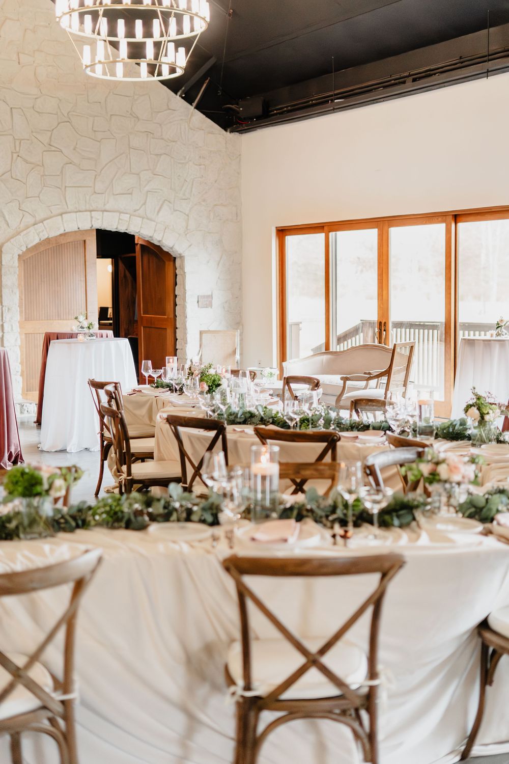 Romantic wedding reception venue with rustic cross-back chairs, floral garland centerpieces, and stone fireplace.