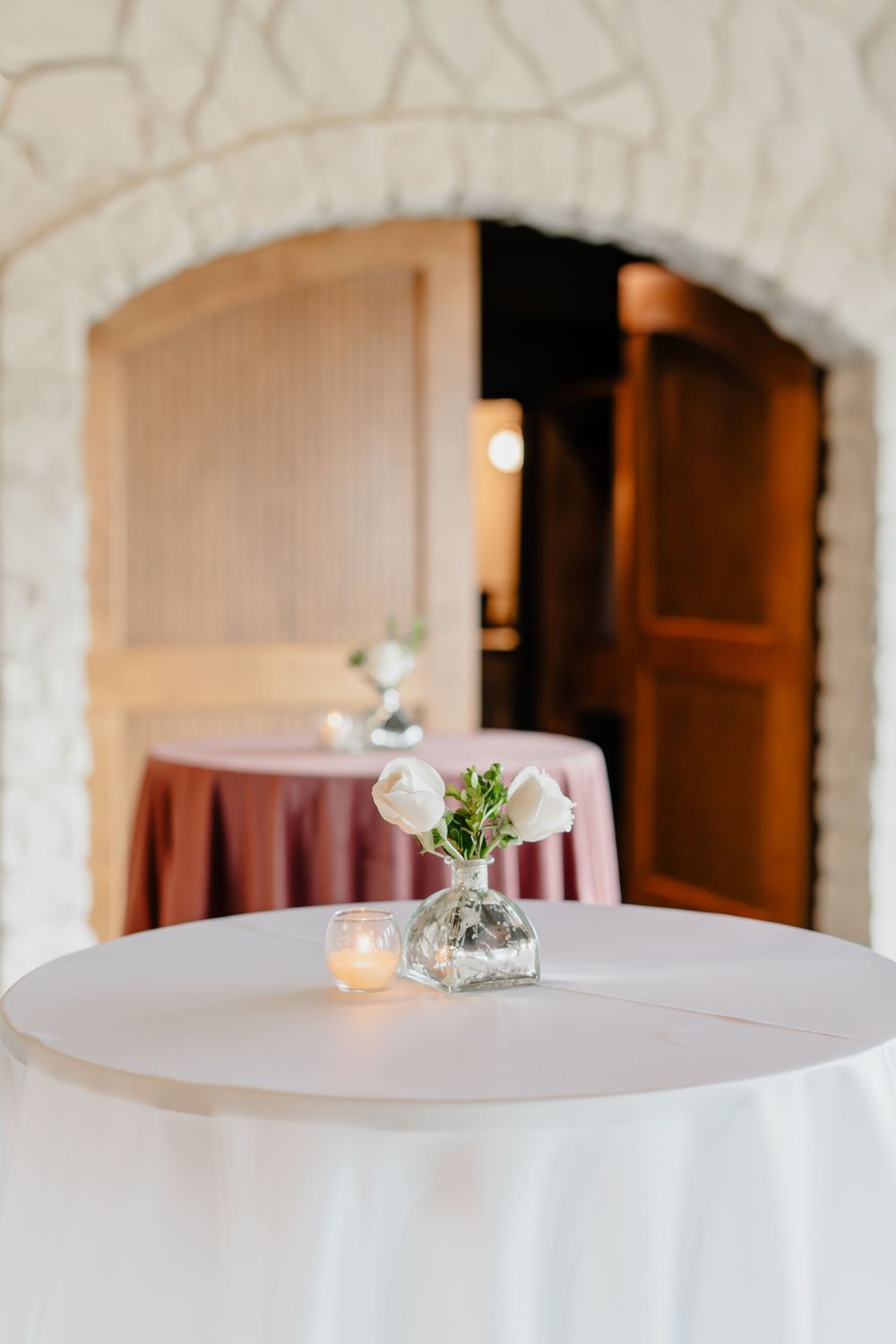 Elegant event table with white linen, glass vase of white flowers, and candle, set against a stone arch doorway.