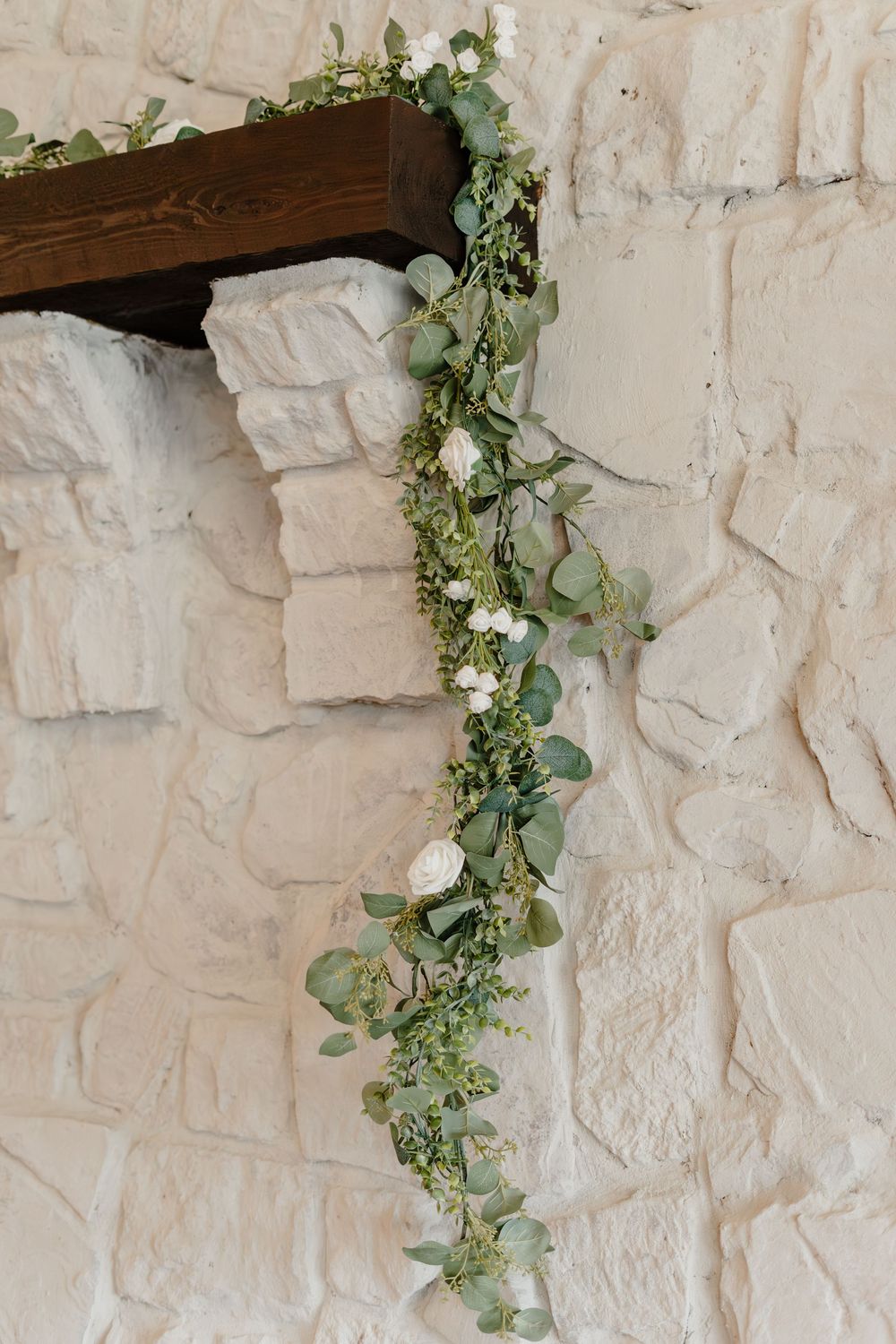 Eucalyptus and white rose garland draped over a rustic wood mantel against a white stone wall.
