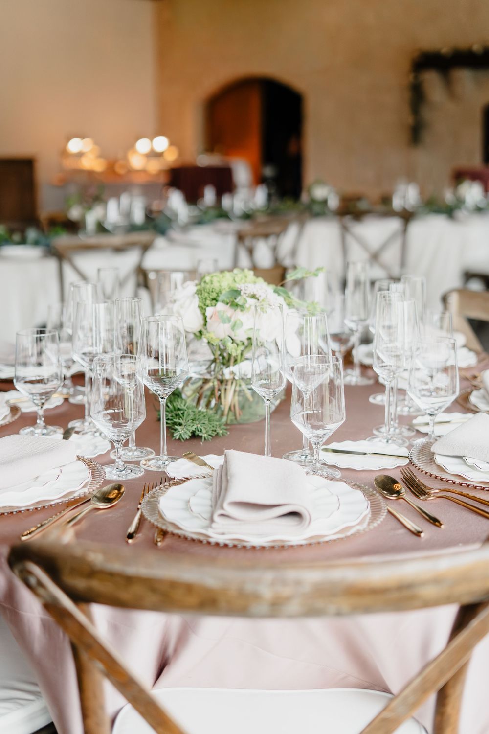 Elegant wedding reception table with blush linens, gold cutlery, crystal glasses, and white floral centerpiece.