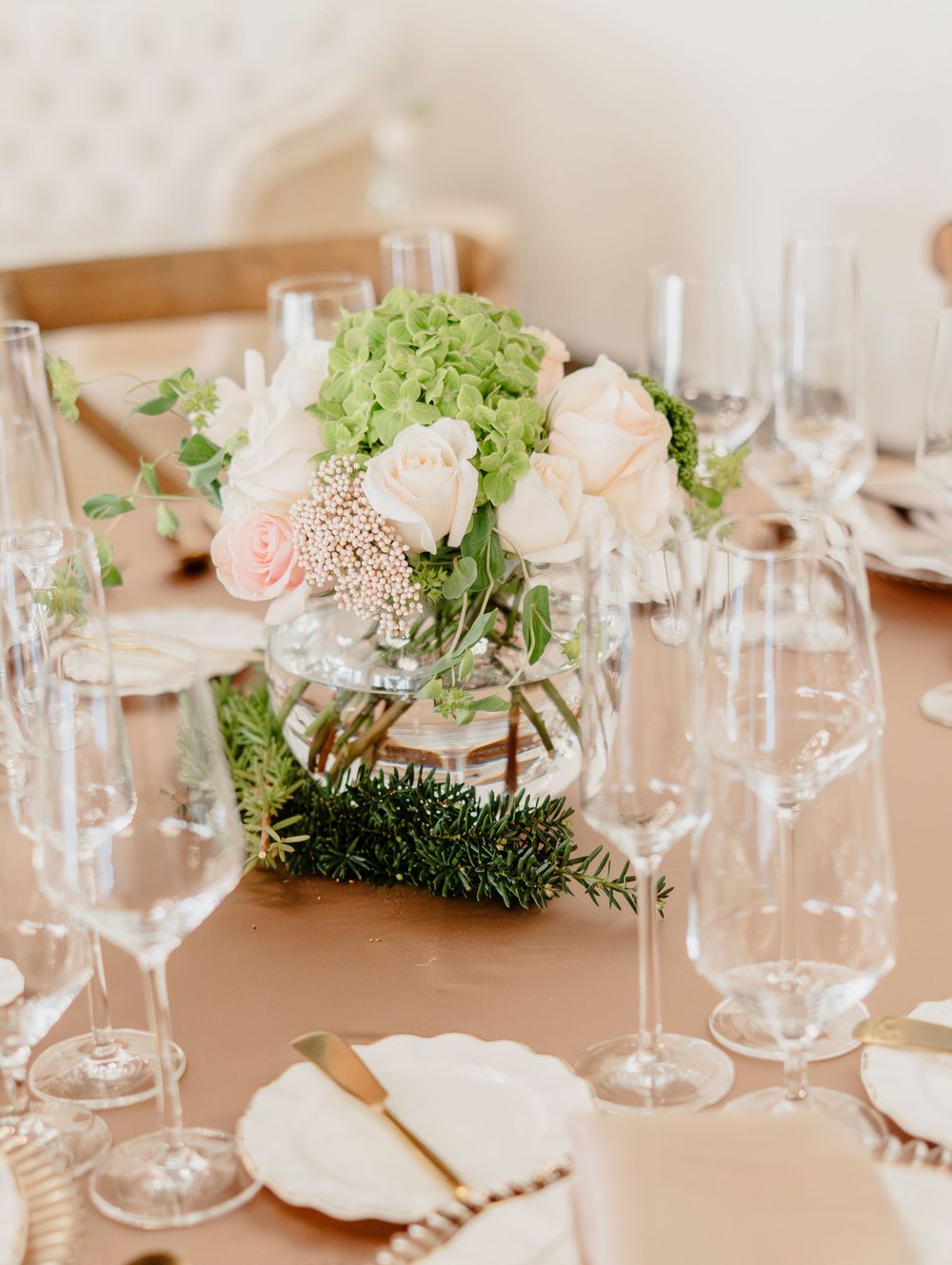 Elegant wedding centerpiece with blush roses and green hydrangeas in a glass vase on a champagne tablecloth.