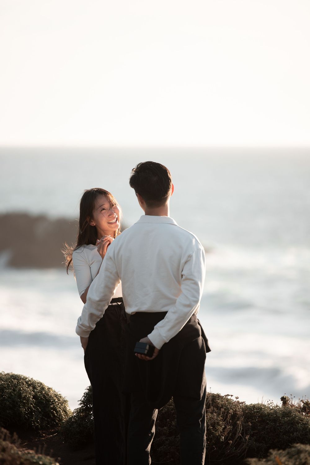 Smiling woman gazes at her partner while standing on rocky coastal cliffs with ocean waves behind them.