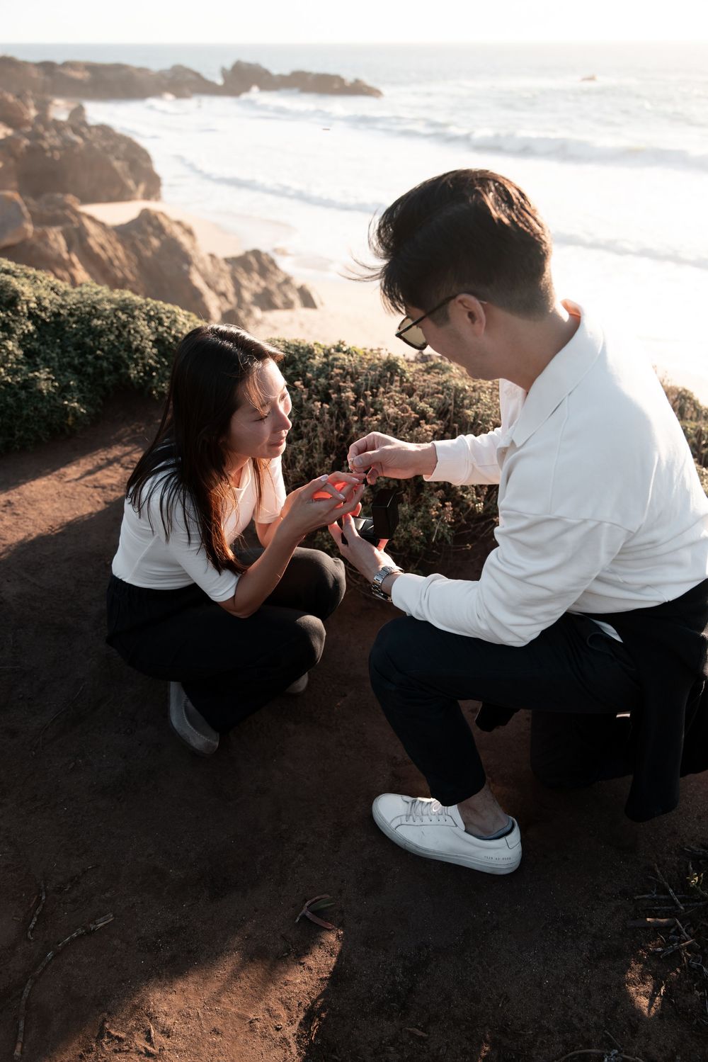 Man proposing to woman on a rocky coastal cliff at sunset, placing a ring on her finger.