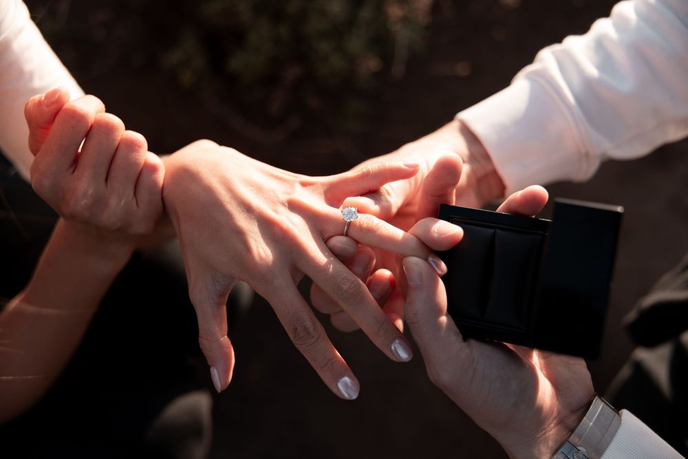 Close-up of a marriage proposal with a diamond engagement ring being placed on a woman's finger outdoors.