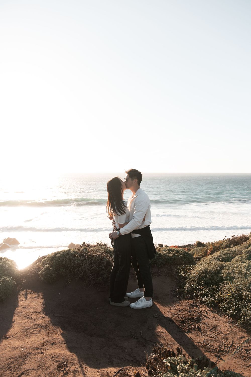 Couple sharing a romantic kiss on a scenic coastal cliff with ocean waves at sunset.