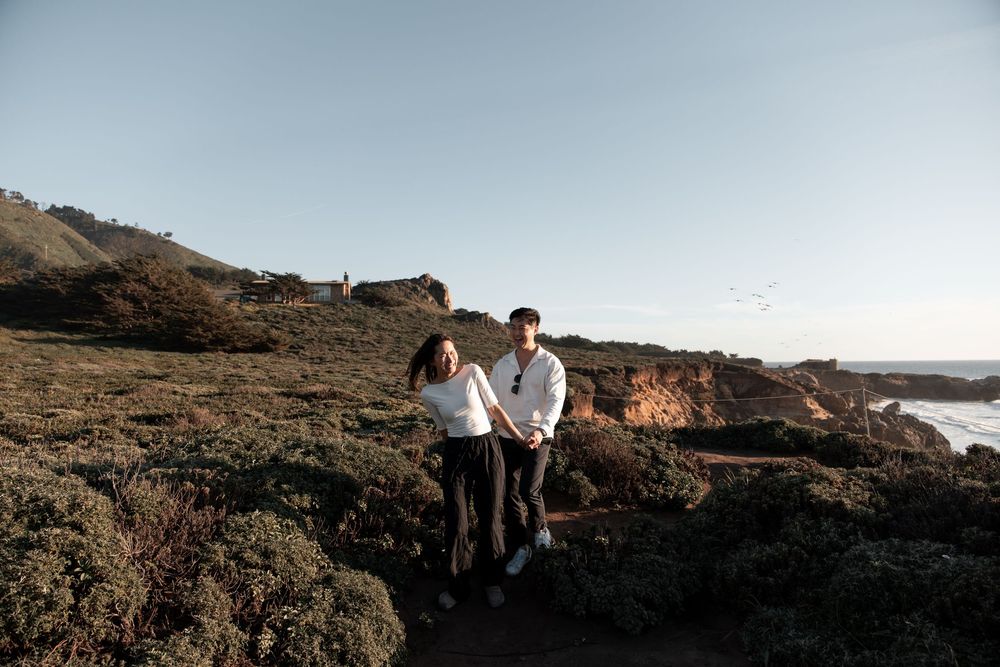 Couple holding hands on rocky coastal trail with ocean views and golden hills at sunset.