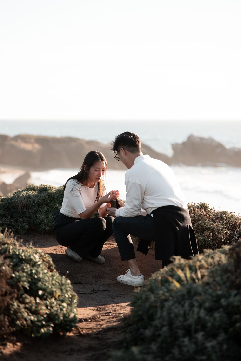Couple sharing a romantic proposal moment on a coastal cliff path surrounded by ocean views.