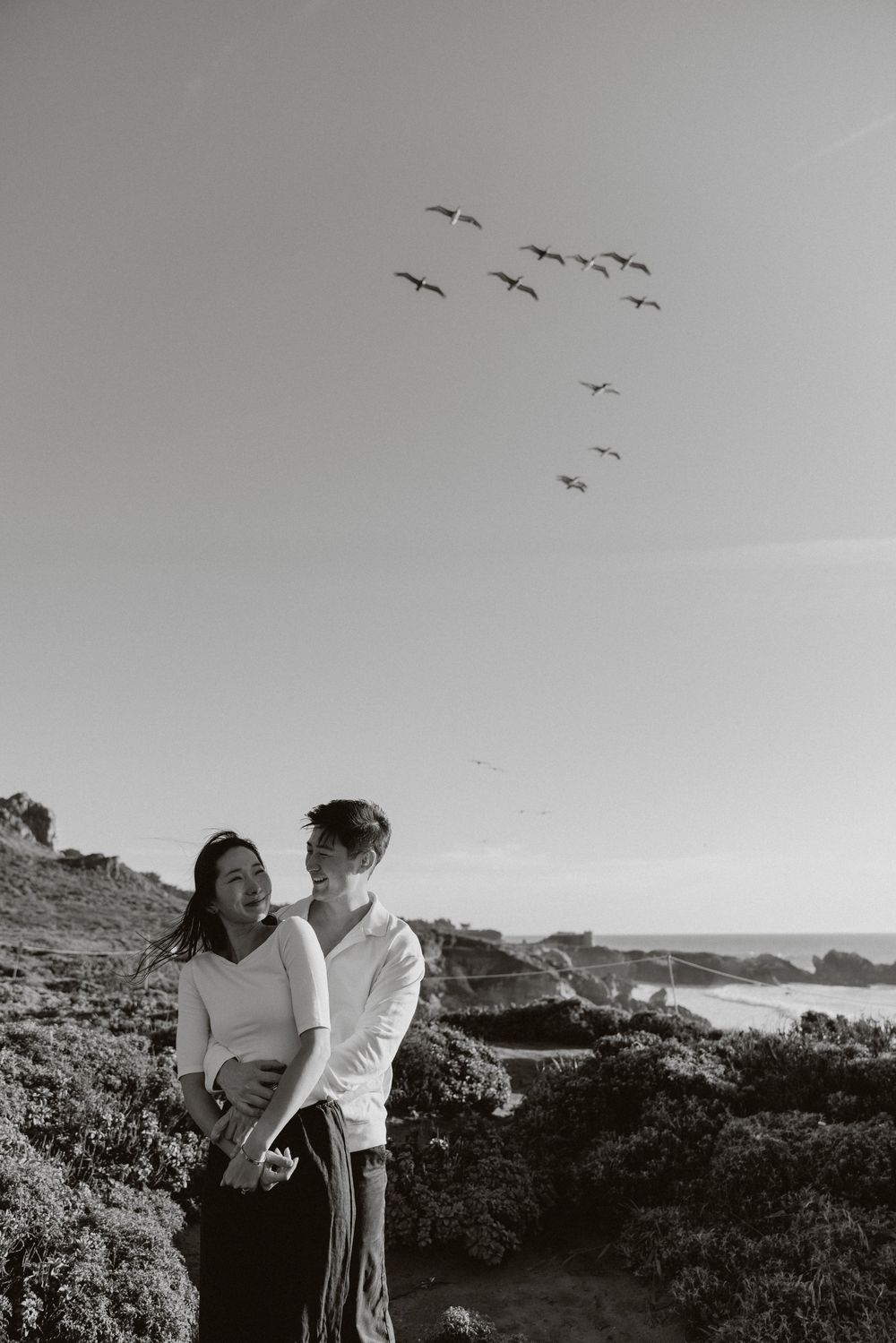 Black and white photo of a couple embracing outdoors near the ocean with birds flying in formation overhead.