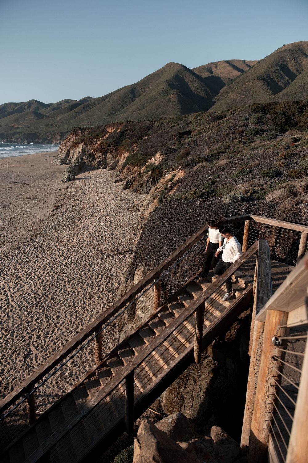 Couple on wooden coastal stairs overlooking sandy beach with rugged green mountains and cliffs along California coastline.