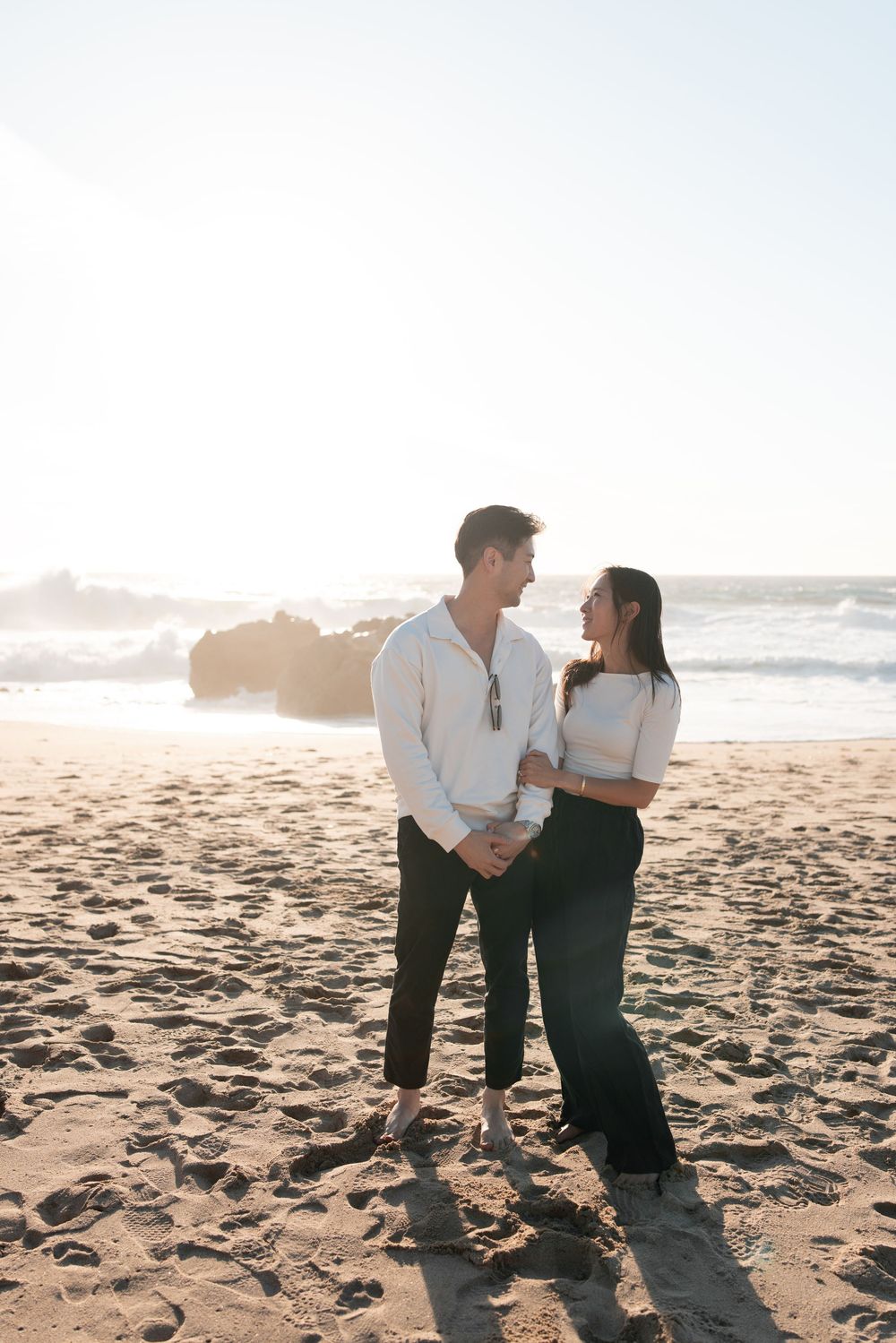 Couple gazing at each other on a sunny beach, man in white shirt and woman in white crop top holding hands.