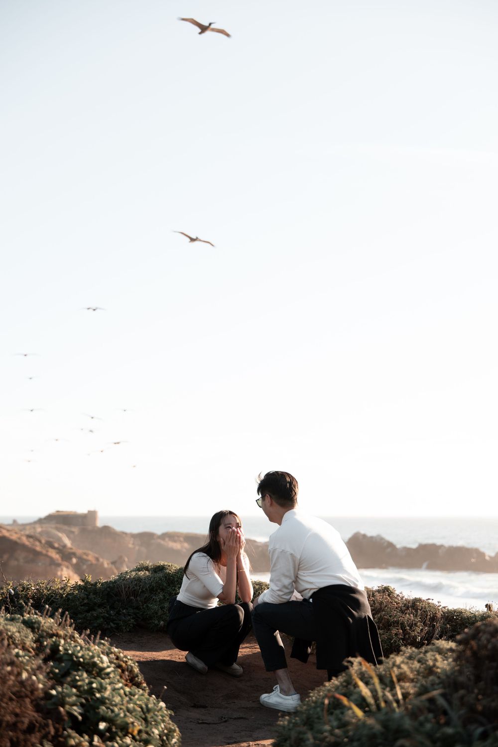 Couple sharing a romantic marriage proposal moment on a coastal cliff at sunset with seagulls flying overhead.