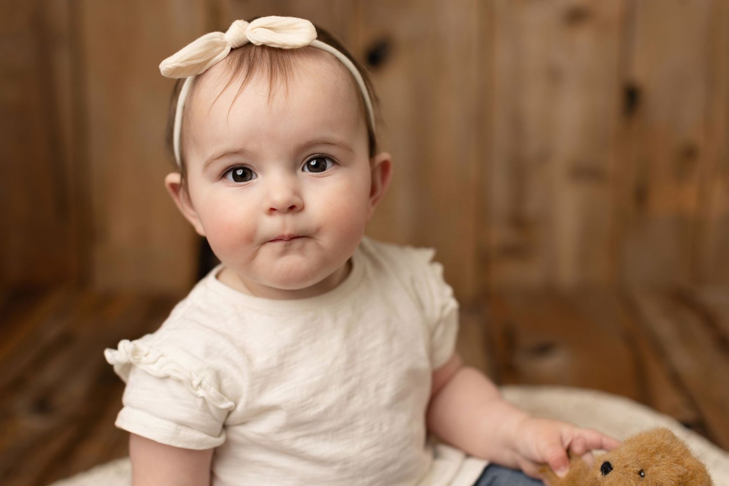 Baby wearing cream-colored outfit and bow headband sits on wooden floor for portrait session. Elizabeth Lang Photography Surrey