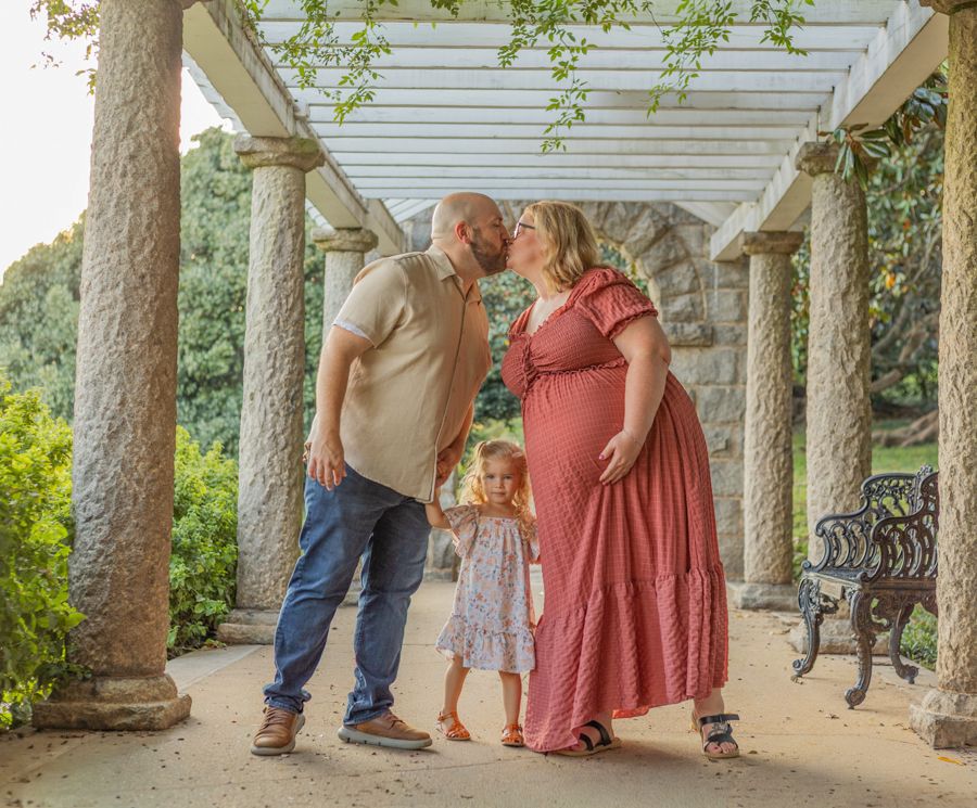 Family maternity photo with couple kissing under pergola columns while young daughter stands between them.