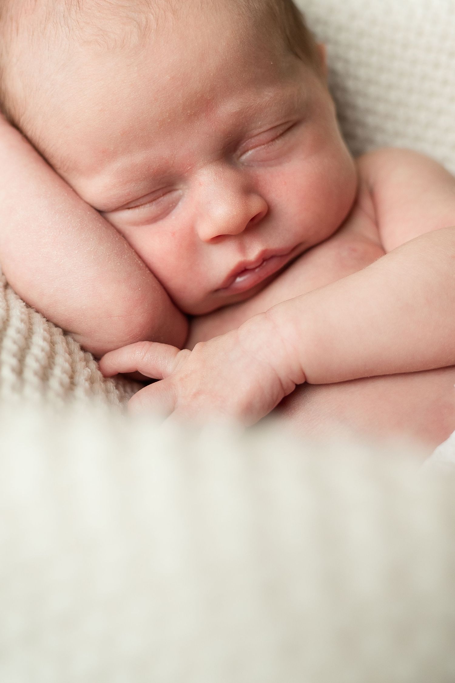 A peaceful sleeping newborn rests on soft white fabric with tiny hands curled up near their face.