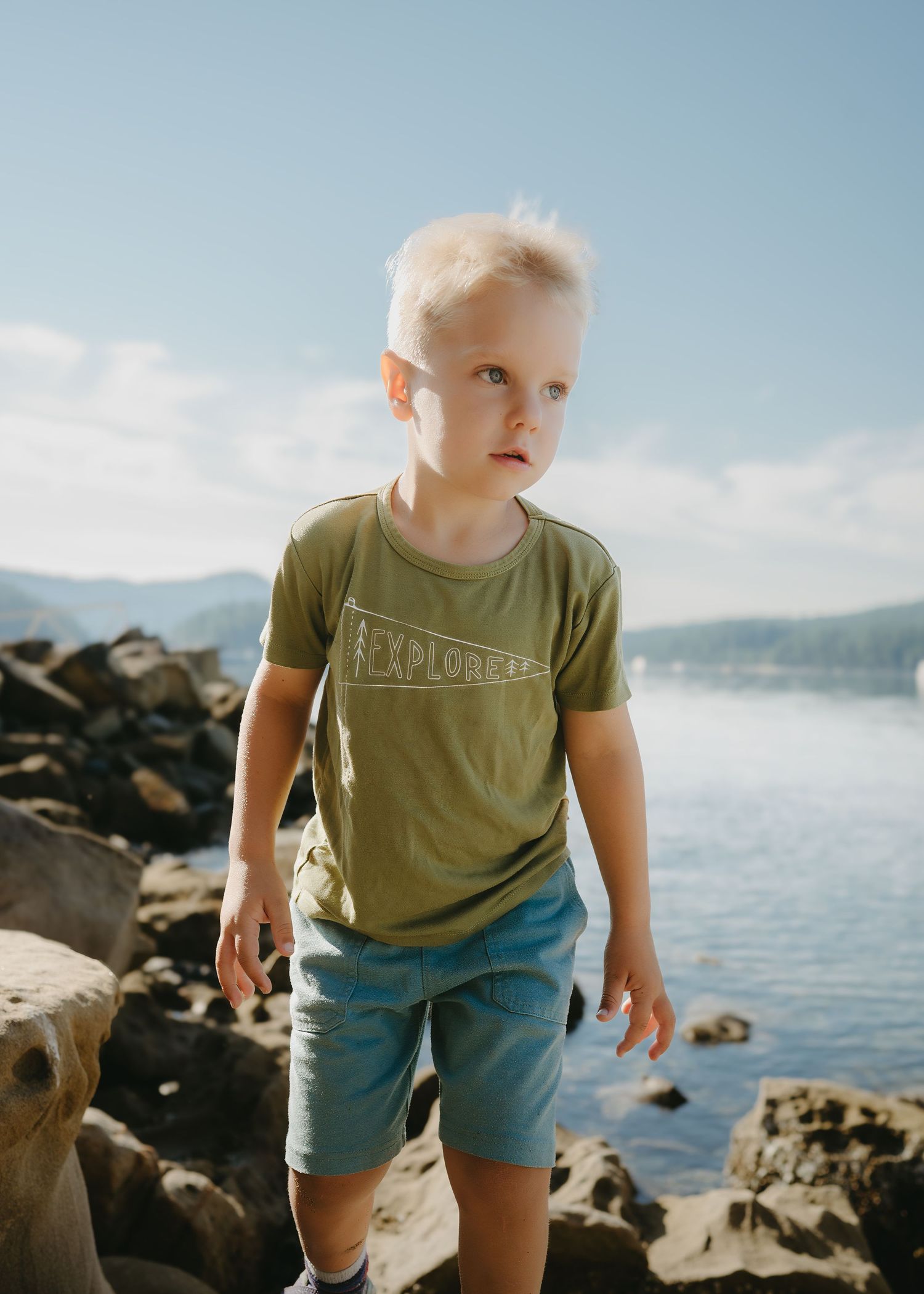 Blonde toddler boy wearing a green Explorer t-shirt and blue shorts standing on rocky shore near a lake.