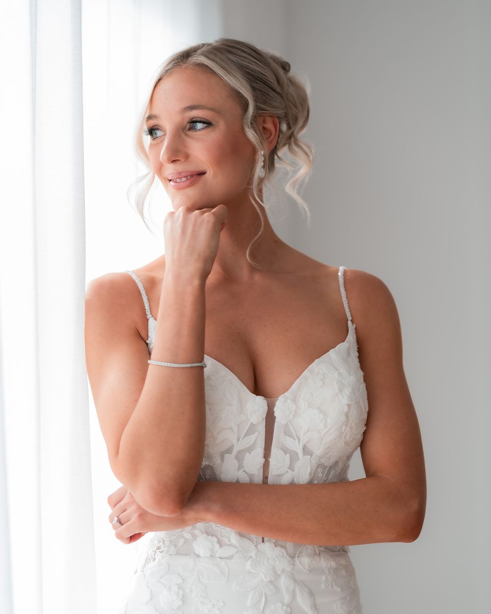 Smiling bride in a white lace spaghetti strap wedding gown with updo hairstyle and delicate bracelet.