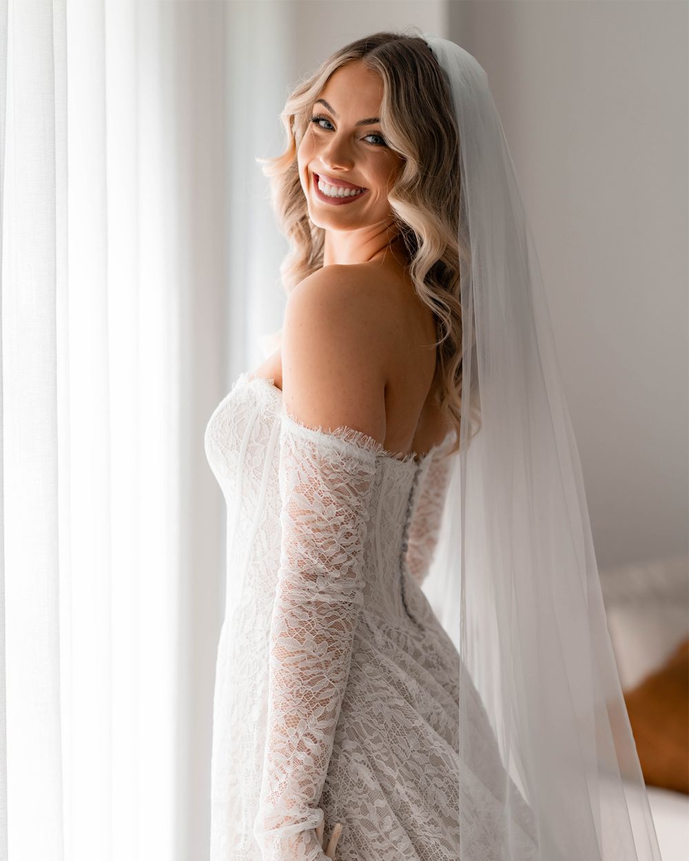 Elegant bride in off-shoulder lace wedding gown with flowing veil poses by window in natural light.