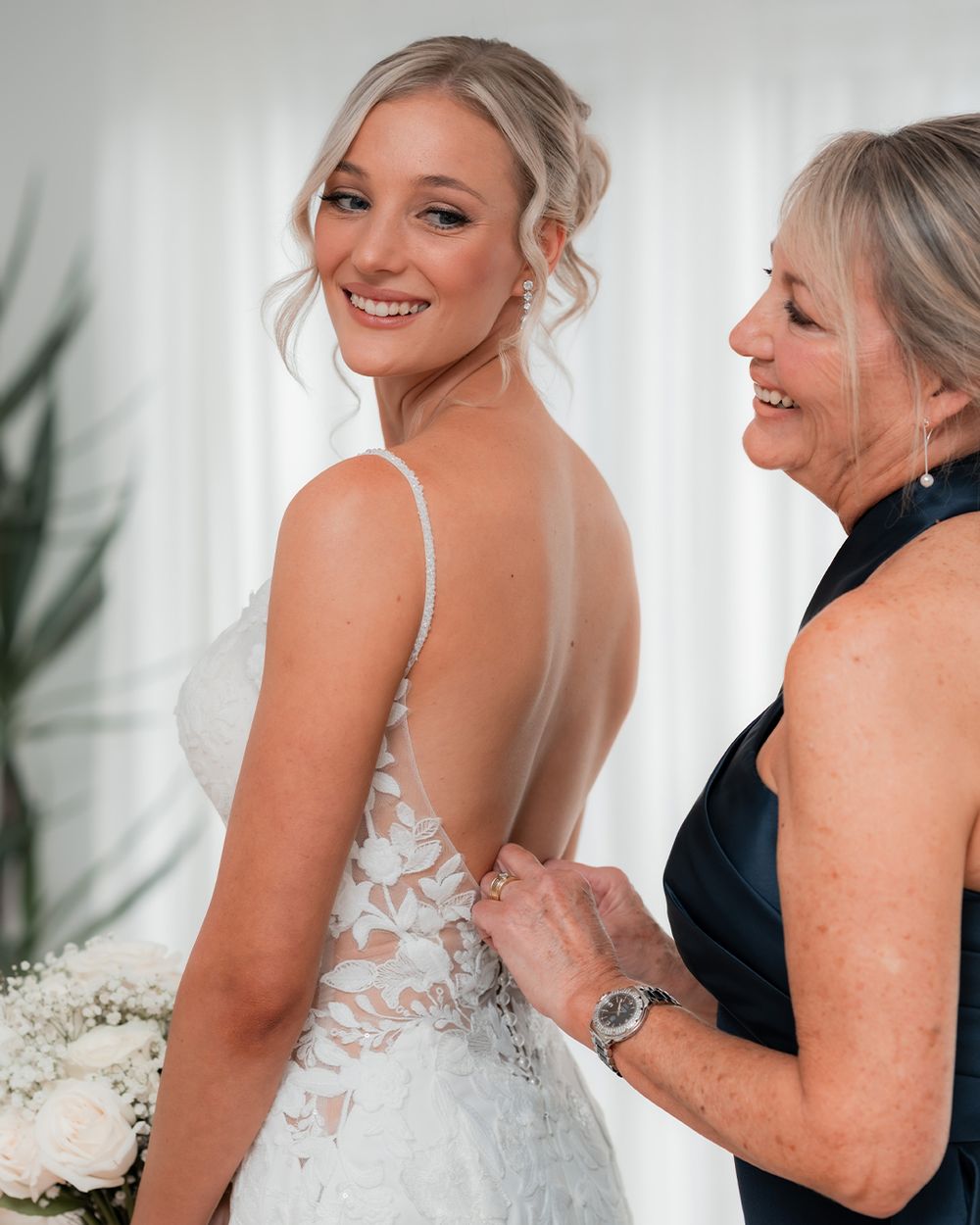 Bride in backless lace wedding gown smiles over shoulder as older woman fastens dress buttons.