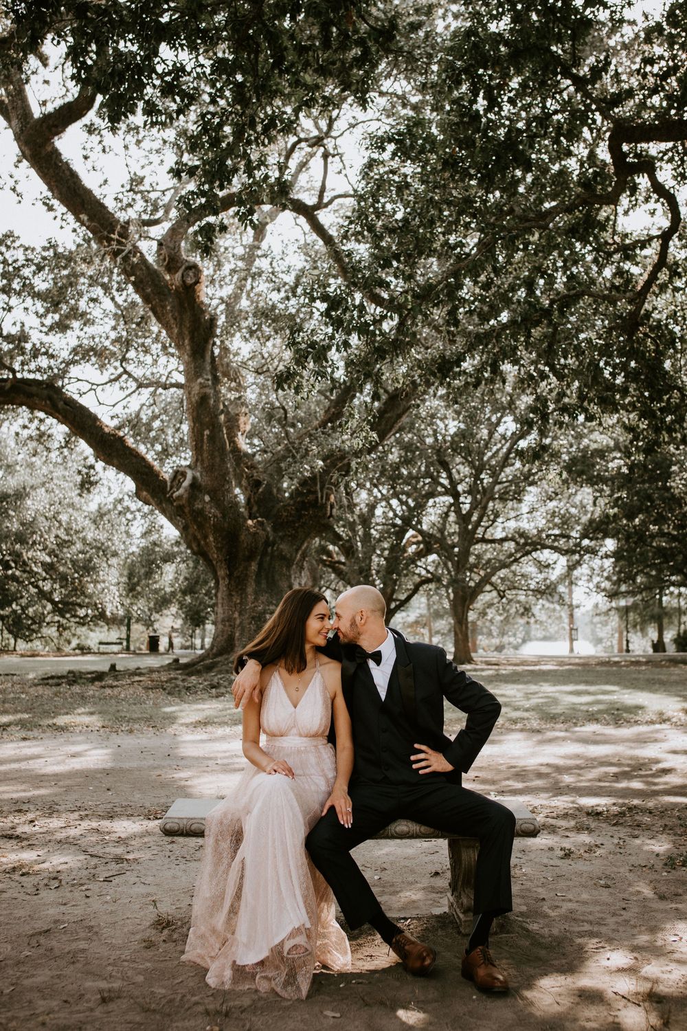 Couple in wedding attire sitting intimately on a stone bench beneath a sprawling oak tree.