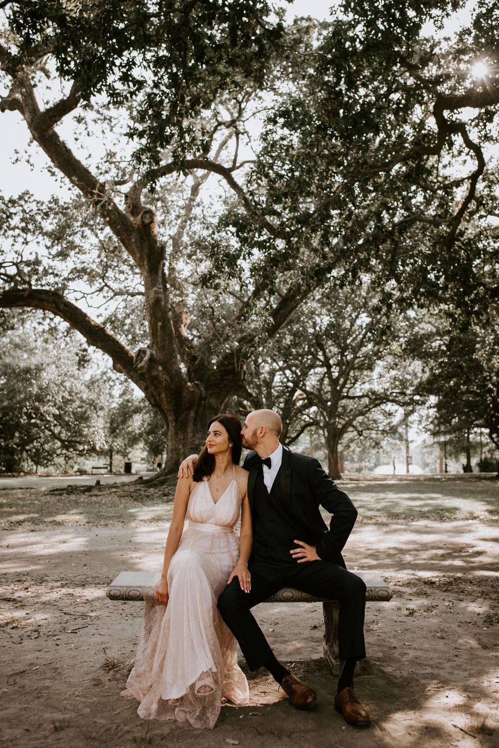 Bride in blush gown and groom in tuxedo share a tender moment on a bench under large oak trees.