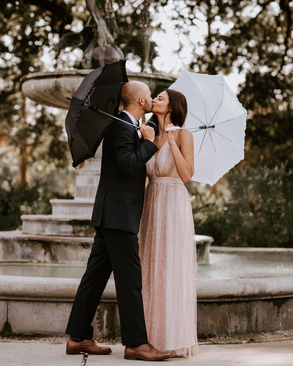 Bride and groom kissing while holding decorative umbrellas near an ornate stone fountain.