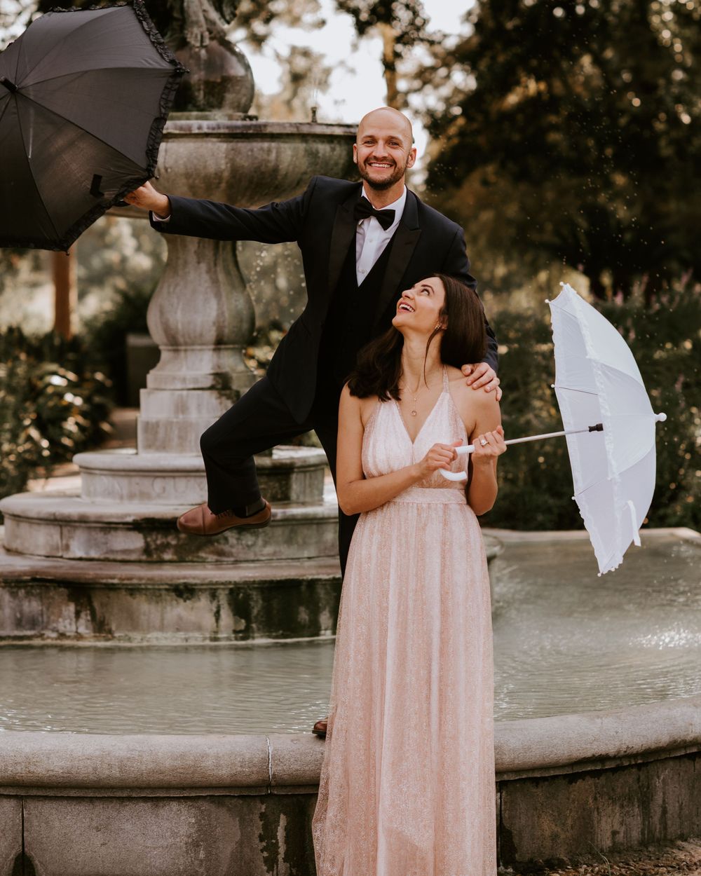 Laughing couple posing playfully with parasols beside a classic stone fountain in a garden.