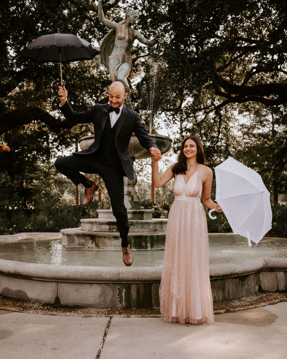 Joyful groom leaps by a fountain as bride holds white umbrella in a lush garden setting.