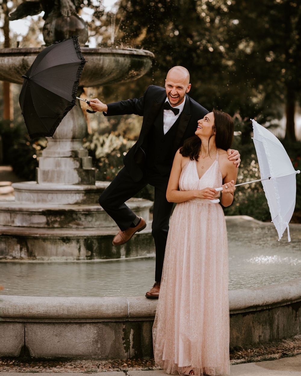 Laughing couple with umbrellas pose playfully by an ornate fountain in a romantic garden.