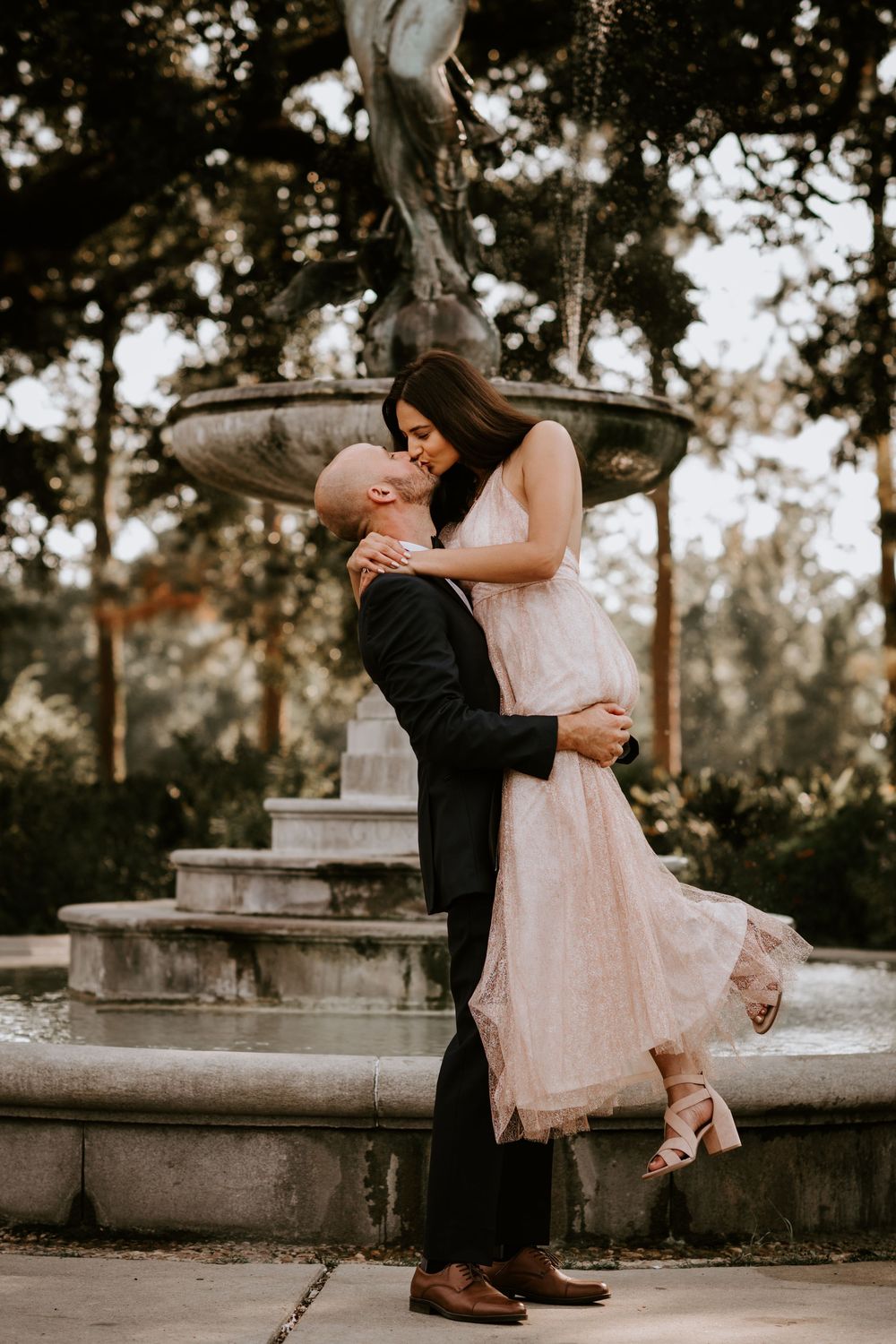 Couple shares a tender kiss as groom lifts bride before a classical stone fountain outdoors.