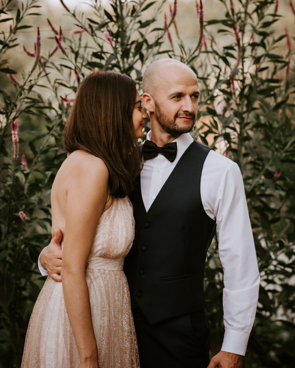 Bride in blush gown embraces bald groom in black vest and bow tie before lush greenery.