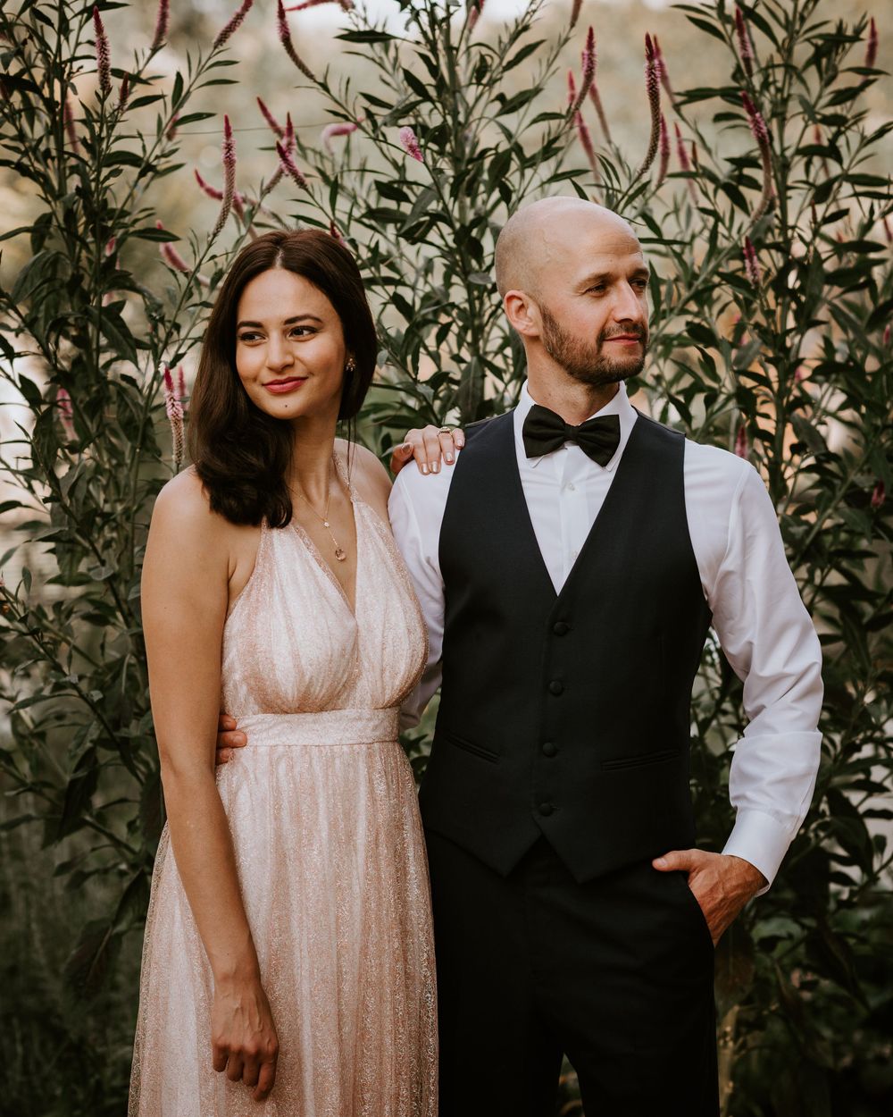 Smiling couple in wedding attire pose together in front of tall green plants outdoors.