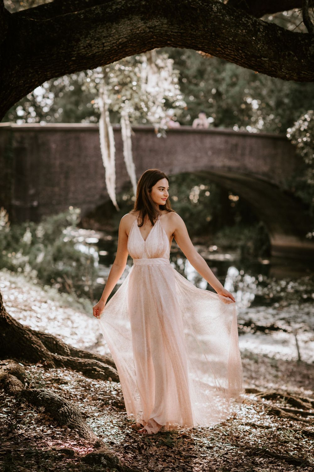 Bride in flowing blush tulle gown twirls gracefully near a stone bridge in a wooded setting.