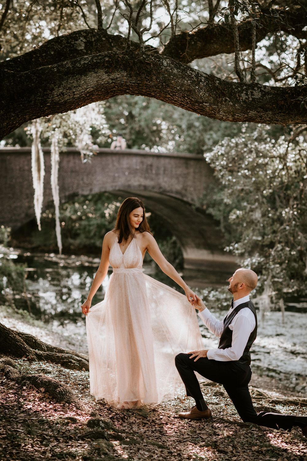Groom kneels holding bride's dress hem beneath a large oak tree near a stone bridge.