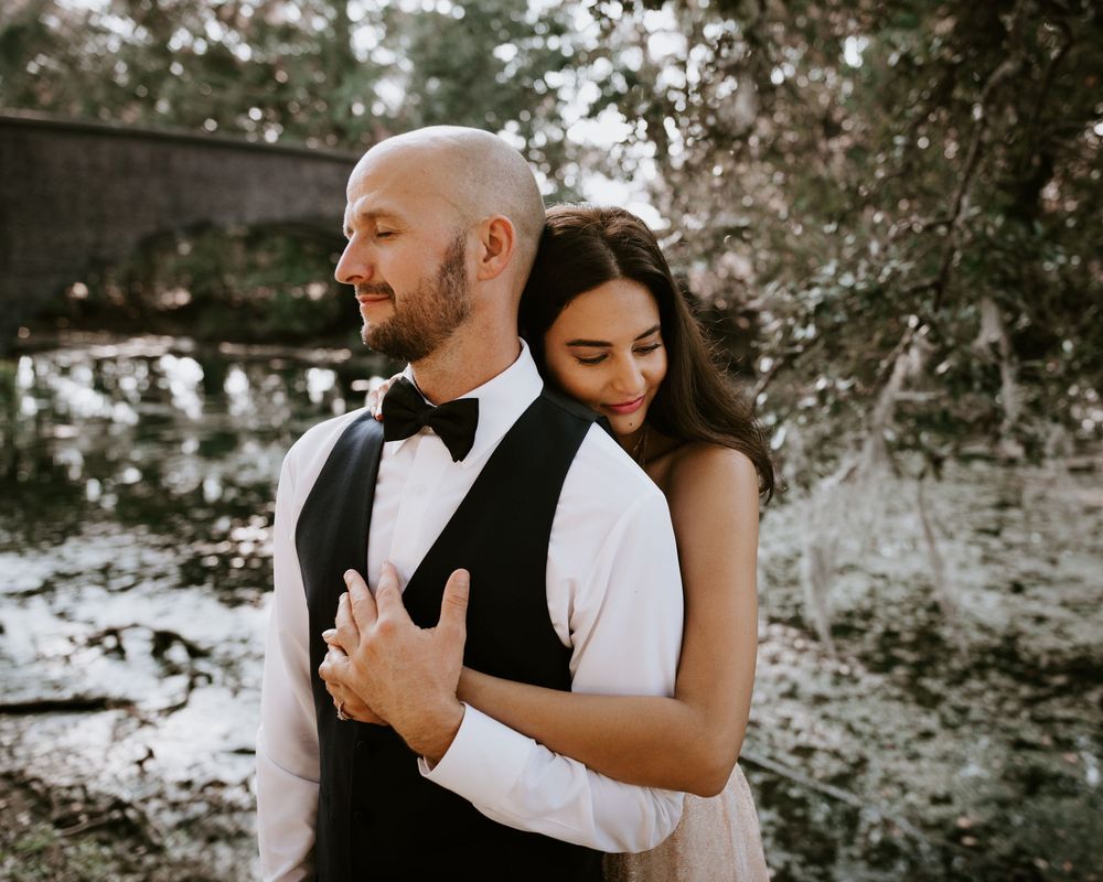Bride embraces groom from behind in a tender moment near a sunlit woodland backdrop.