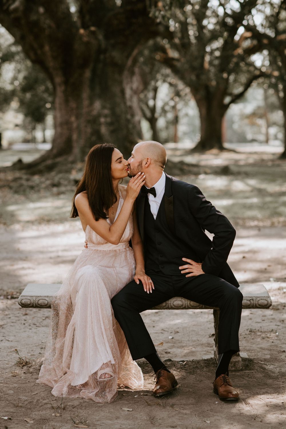 Bride and groom sharing a romantic kiss while seated on a stone bench in a park setting.