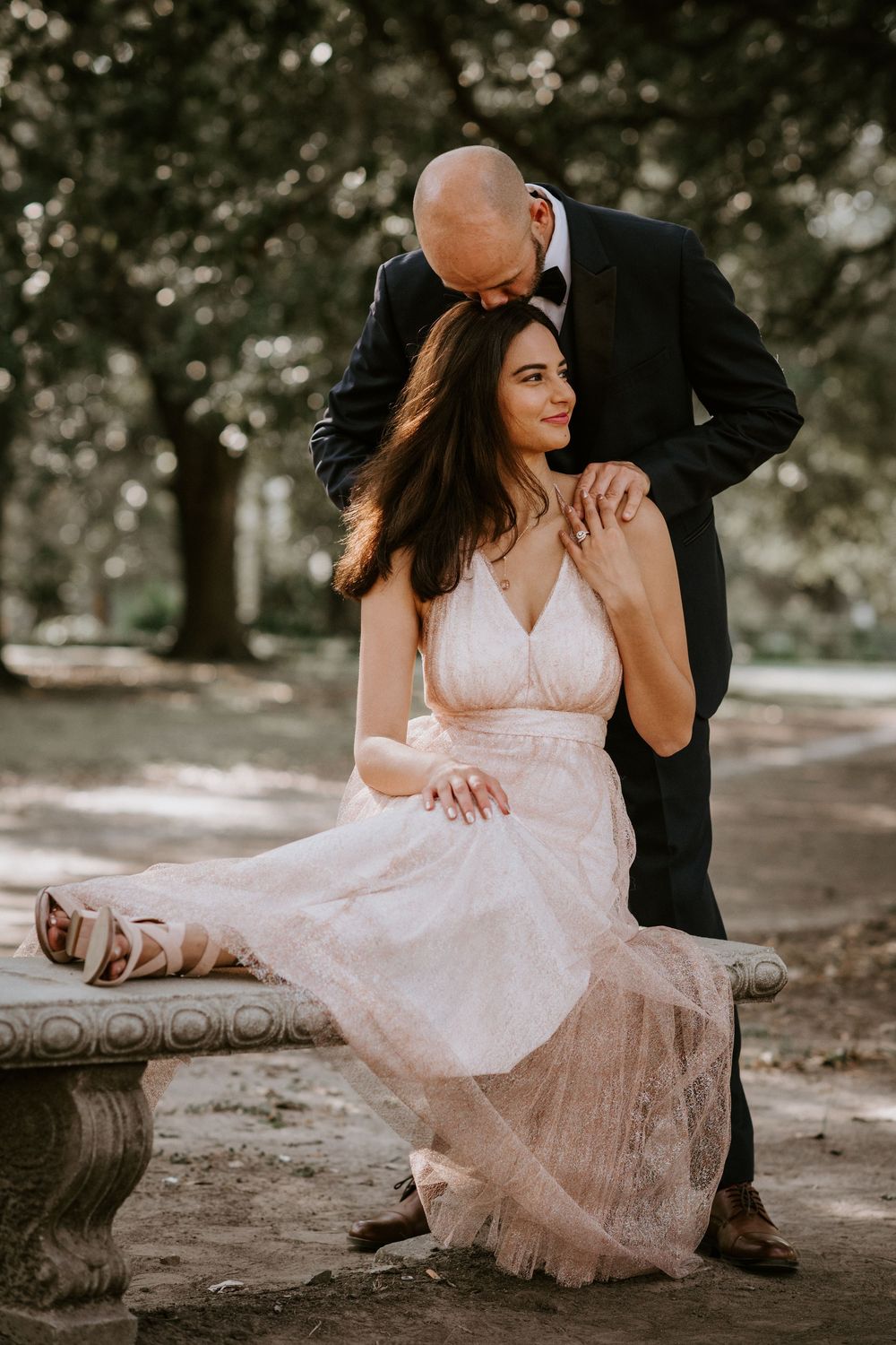 Groom in black tuxedo leans over smiling bride in blush lace gown seated on a stone bench.