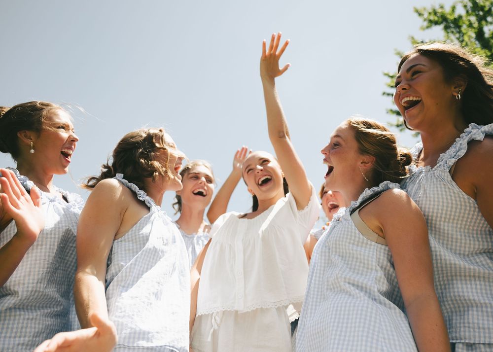 Joyful bride in white dress celebrating with bridesmaids in blue gingham dresses outdoors on a sunny day.