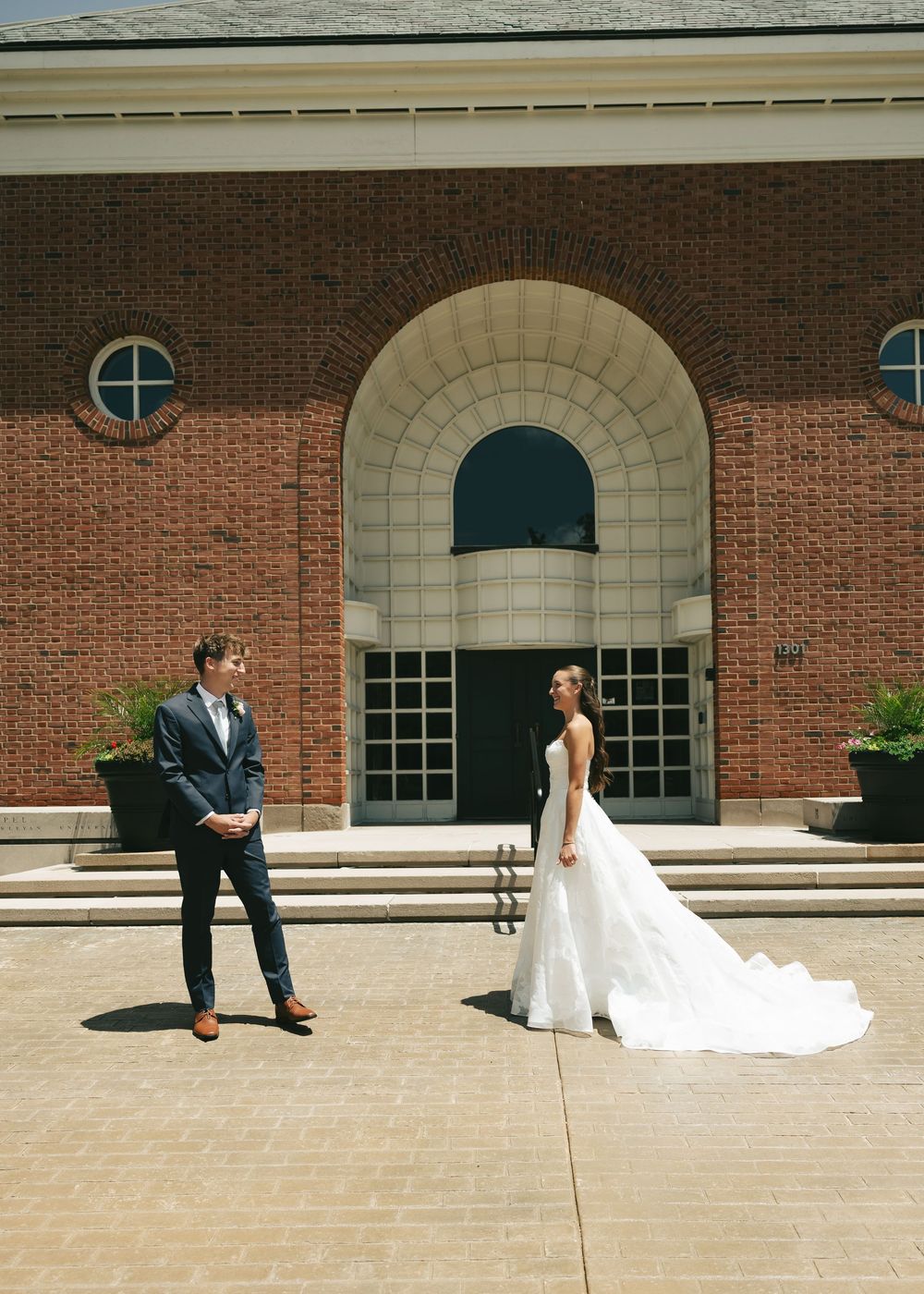 Bride in white gown and groom in navy suit share a first look moment outside a brick building on their wedding day.