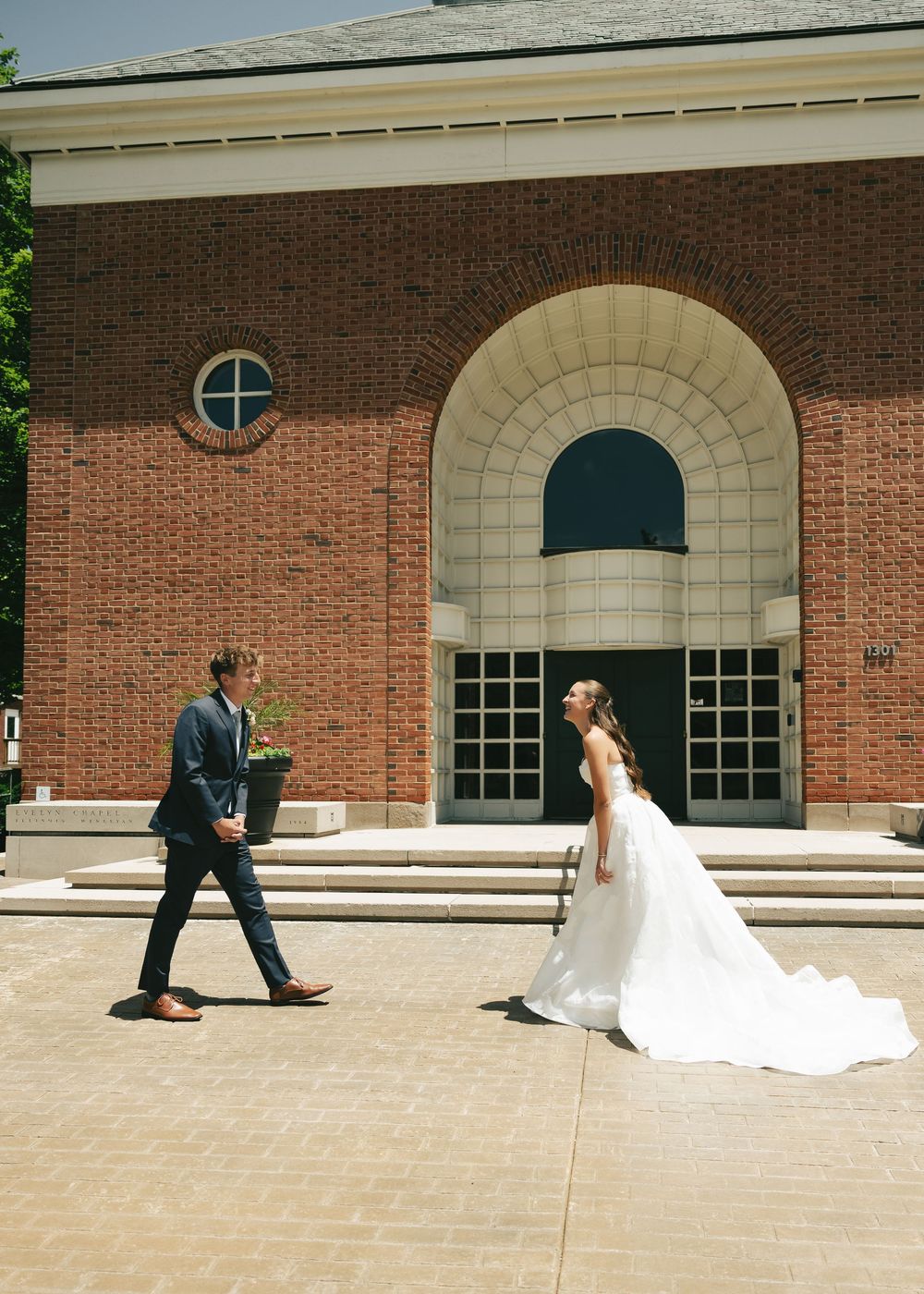 Bride in white gown and groom in navy suit walk toward each other outside a brick building on their wedding day.