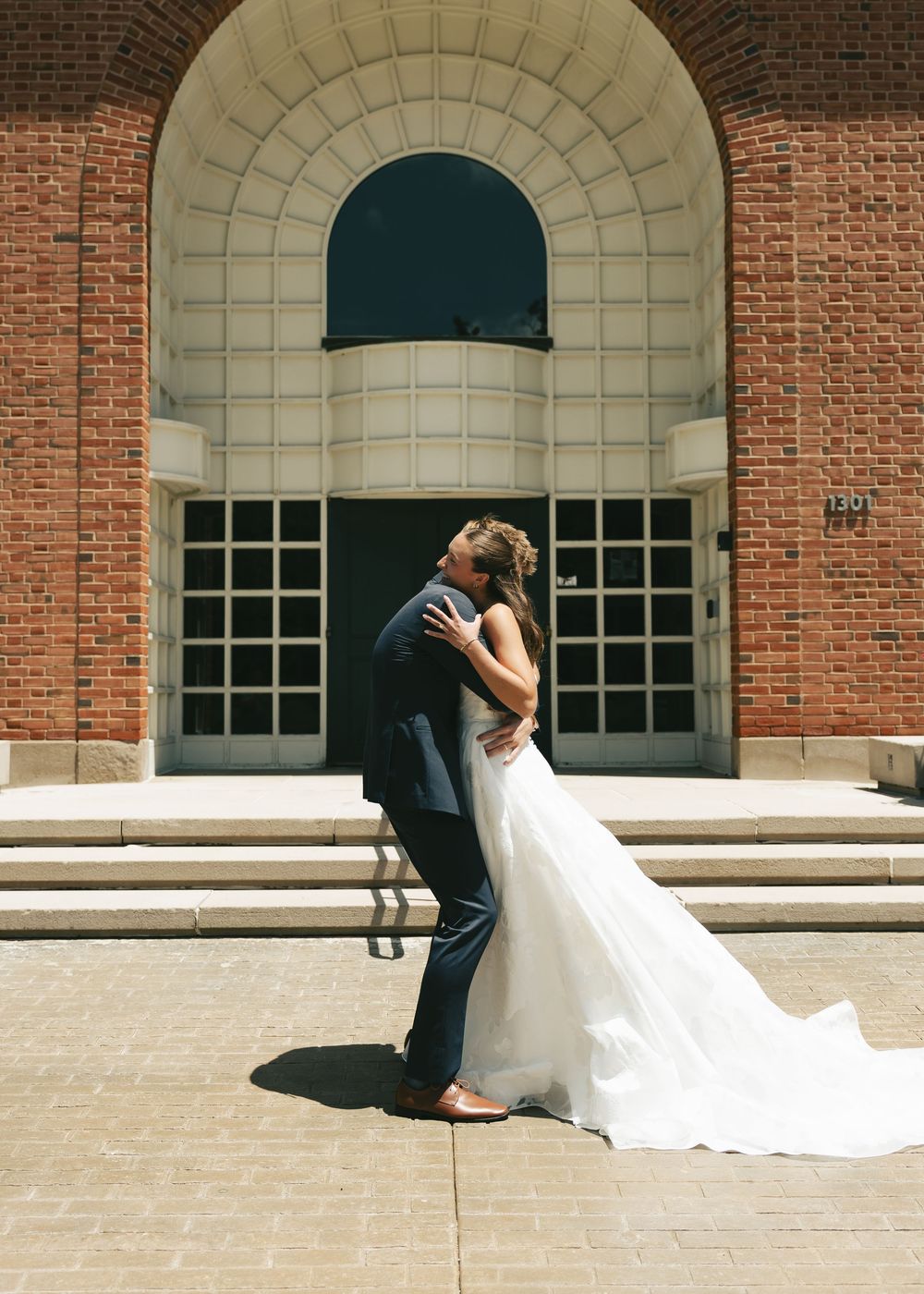 Bride and groom share romantic kiss in front of brick arch building, bride in flowing white gown, groom in navy suit.