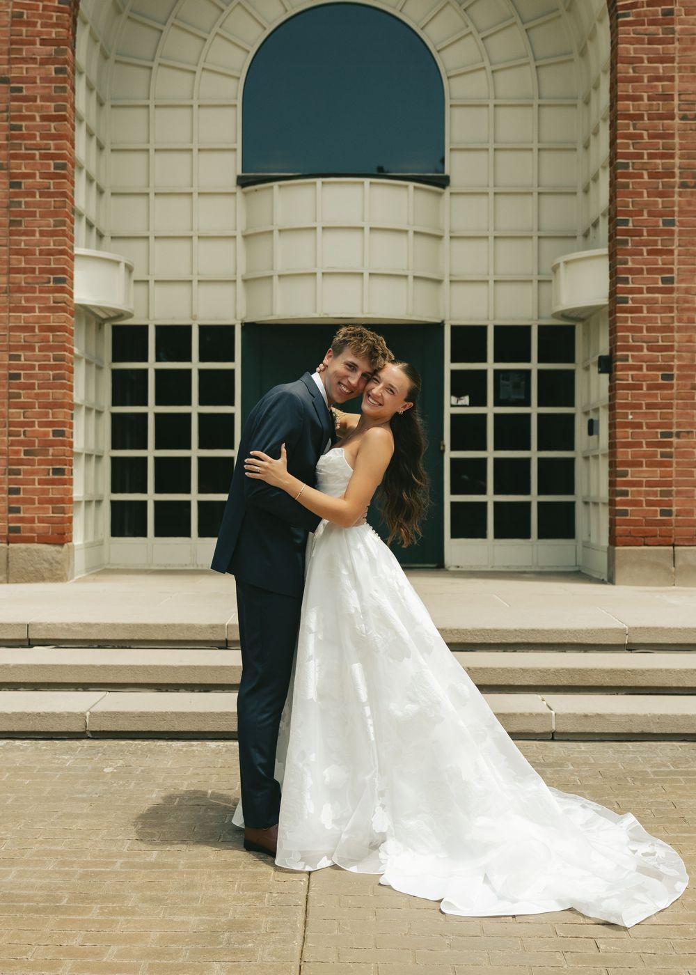 Happy newlyweds embrace in front of an arched brick building, bride in white gown and groom in navy suit.