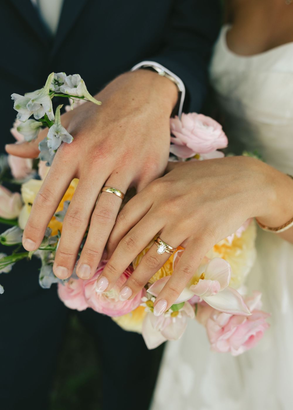 Newlywed couple displaying gold wedding rings on hands resting over a bridal bouquet of pink and blue flowers.