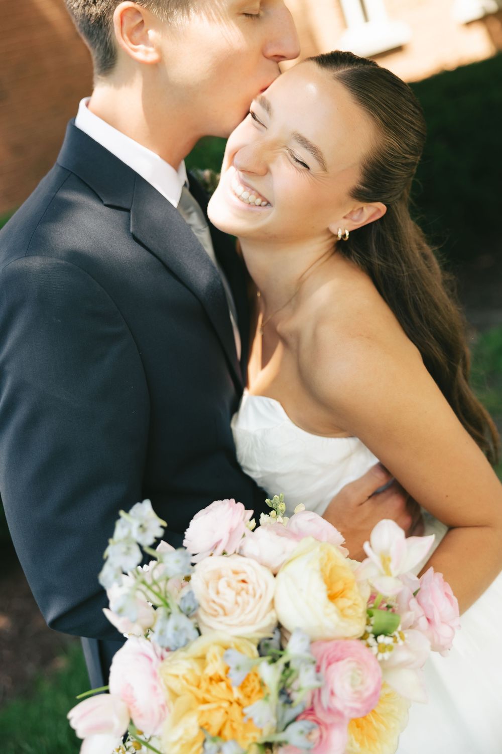 Bride smiles joyfully as groom kisses her forehead, holding a pastel bouquet of pink and yellow roses.